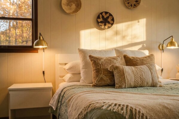 A bedroom with a bed, pillows, bedside lamps, a nightstand, and wall decor. - Summer Oasis: Hot Tub, Huge Yard, Pool + Lakes (Long Pond)