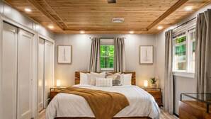 A bedroom with a wooden ceiling, a large bed, bedside tables, a window with curtains, and framed pictures on the wall. - Hot Tub | Game Room | Fire Pit | Family Friendly (Albrightsville)