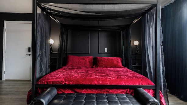 A four-poster bed with red bedding, black headboard, and curtains, in a room with a door and wall-mounted lights. - Lovely 1-bedroom studio with AC in Phoenix (Phoenix)
