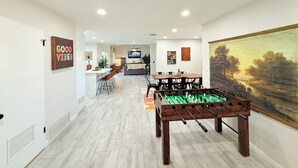 A modern living room with a foosball table, a dining area with a painting, and a TV in the background. - 4BR Family & group Oasis in Mesa – Big Backyard & Steps from Sloan Park & ASU!! (Mesa)