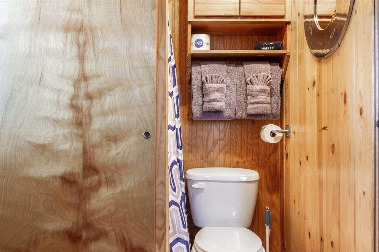 A small wooden bathroom with a toilet, a shelf with towels, and a makeup kit.
