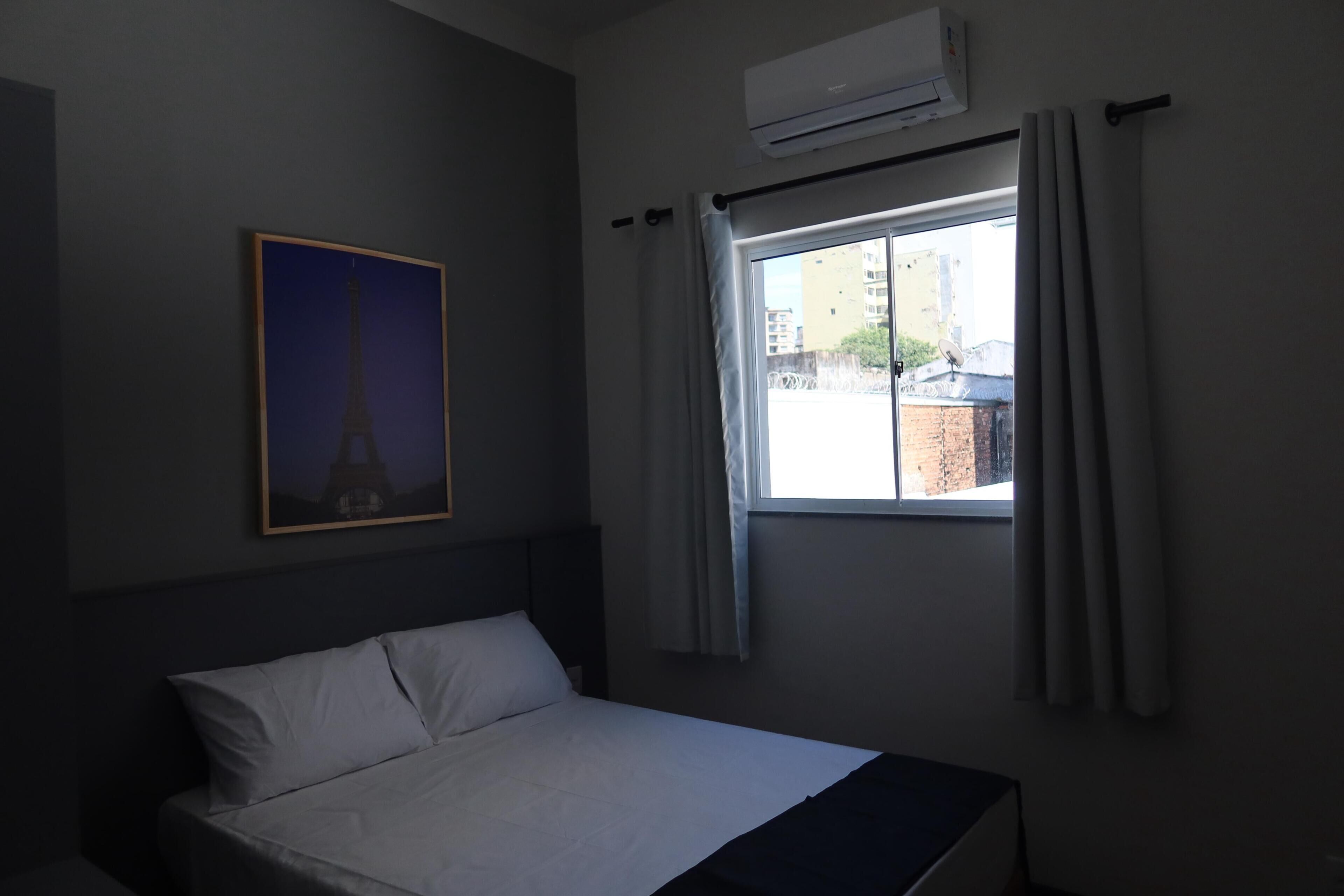 Quarto de hotel com cama, janela com cortinas, ar condicionado e uma fotografia emoldurada da Torre Eiffel na parede.