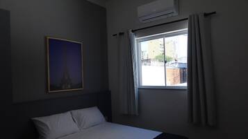 Quarto de hotel com cama, janela com cortinas, ar condicionado e uma fotografia emoldurada da Torre Eiffel na parede.