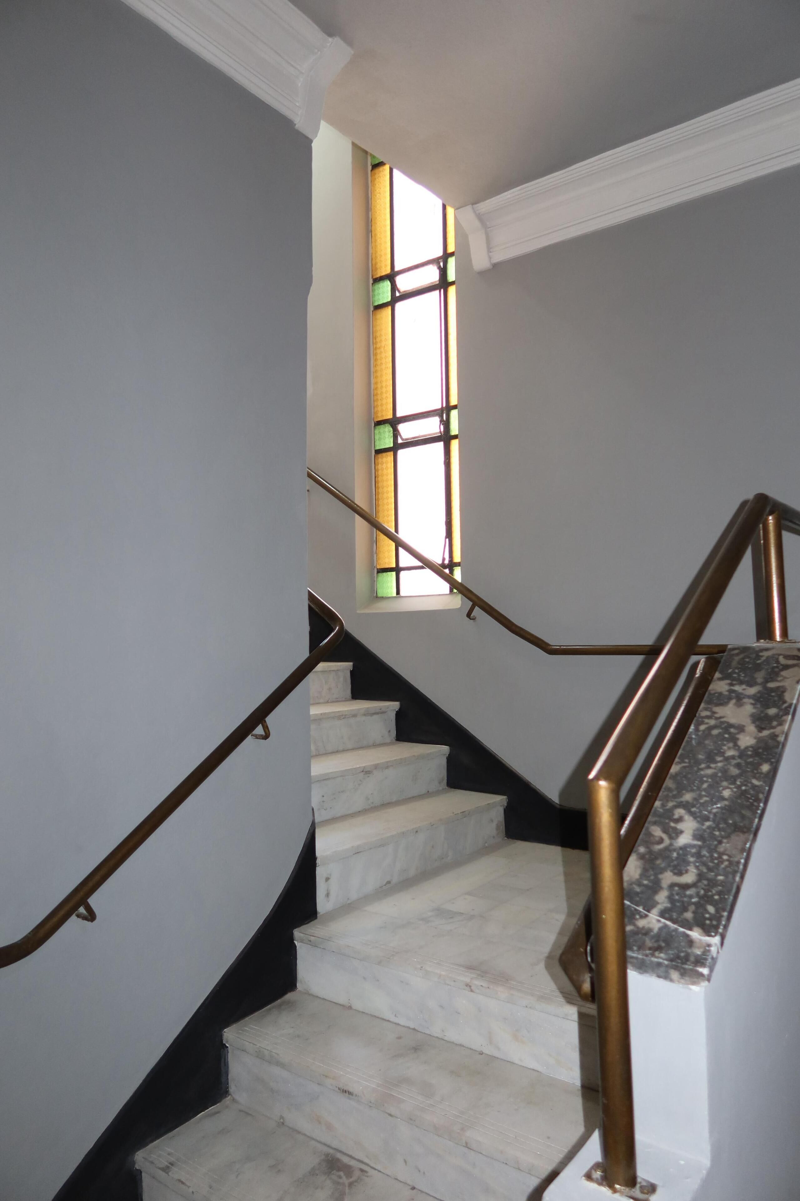A staircase with marble steps and brass handrails, leading to a room with a window featuring stained glass.