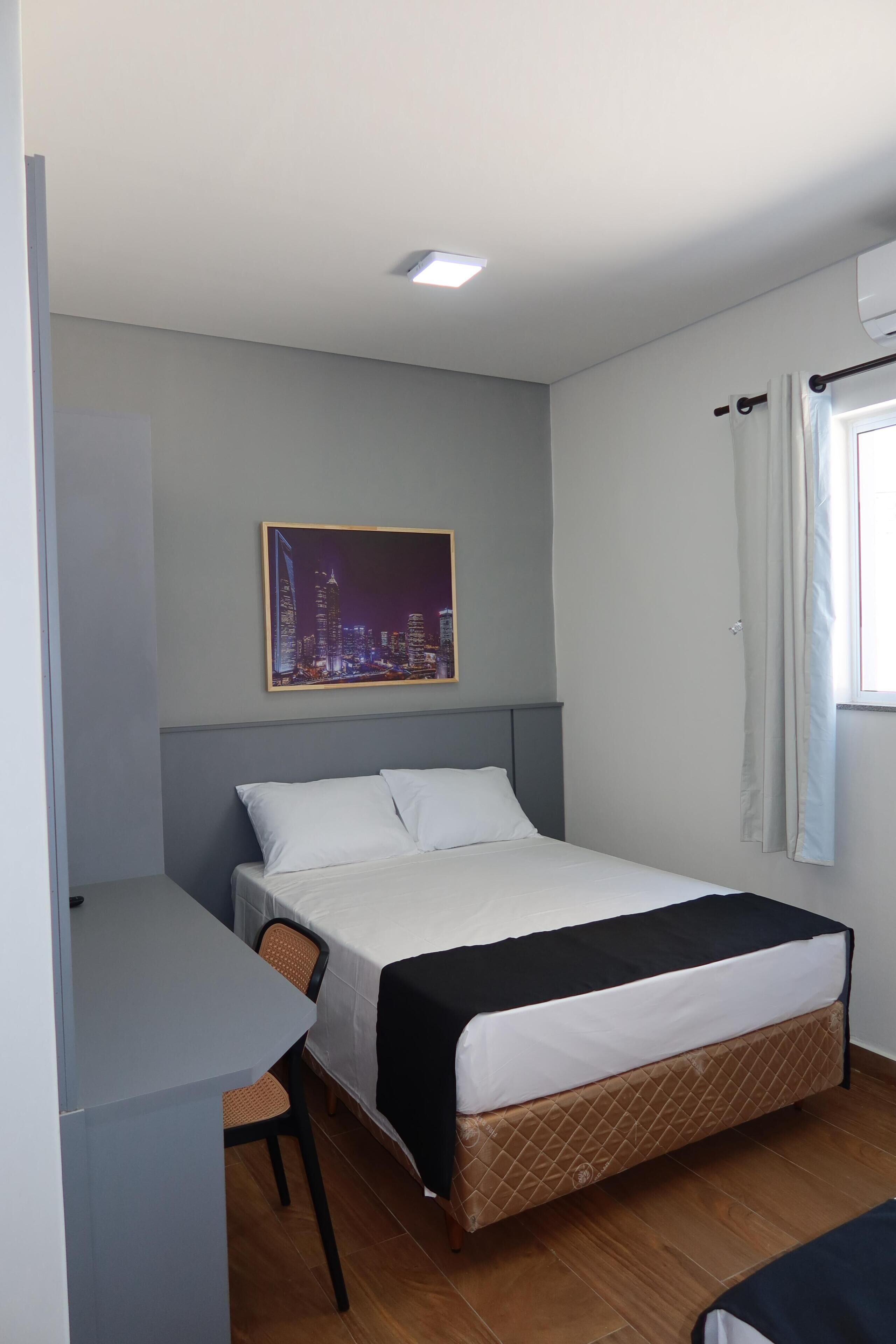 A hotel room with a bed, a desk, a chair, a cityscape painting on the wall, and a window with curtains.