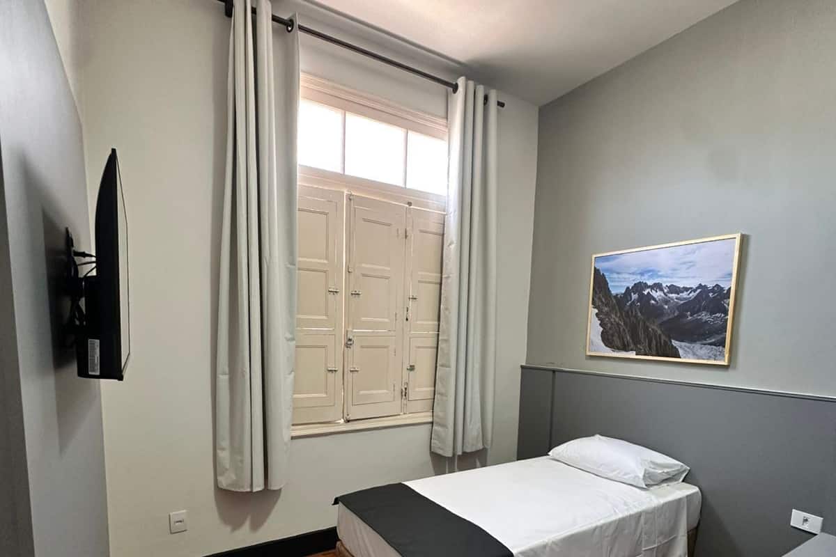 Une chambre à coucher comprenant un lit, un bureau, une chaise, une fenêtre avec des rideaux, un téléviseur fixé au mur et un tableau accroché au mur.