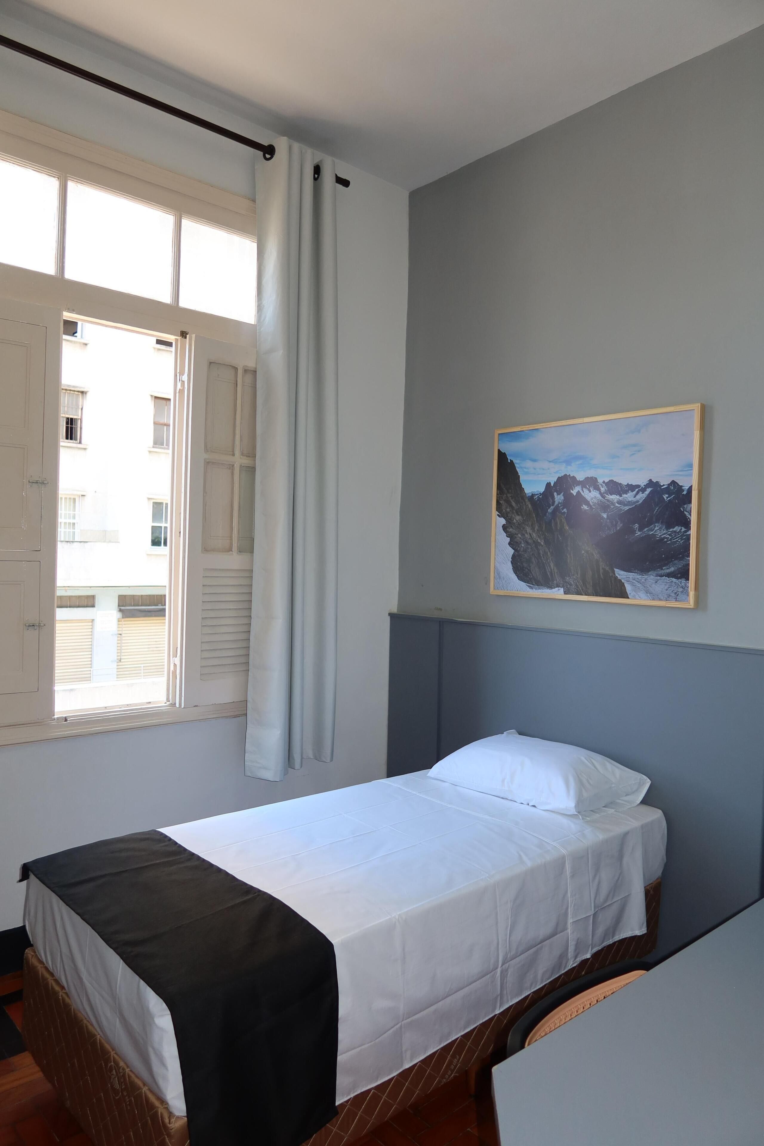 A single bed with a white and black bedspread, a window with curtains, and a framed picture of mountains on the wall.