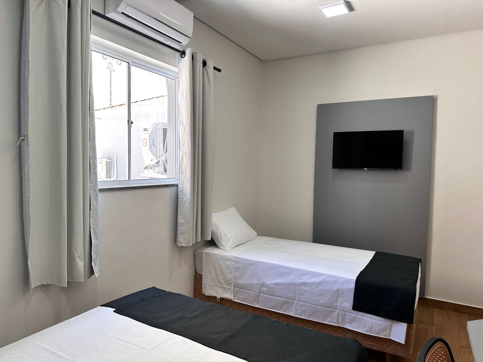 Una habitación con dos camas, una ventana con cortinas, un televisor montado y una lámpara en el techo.
