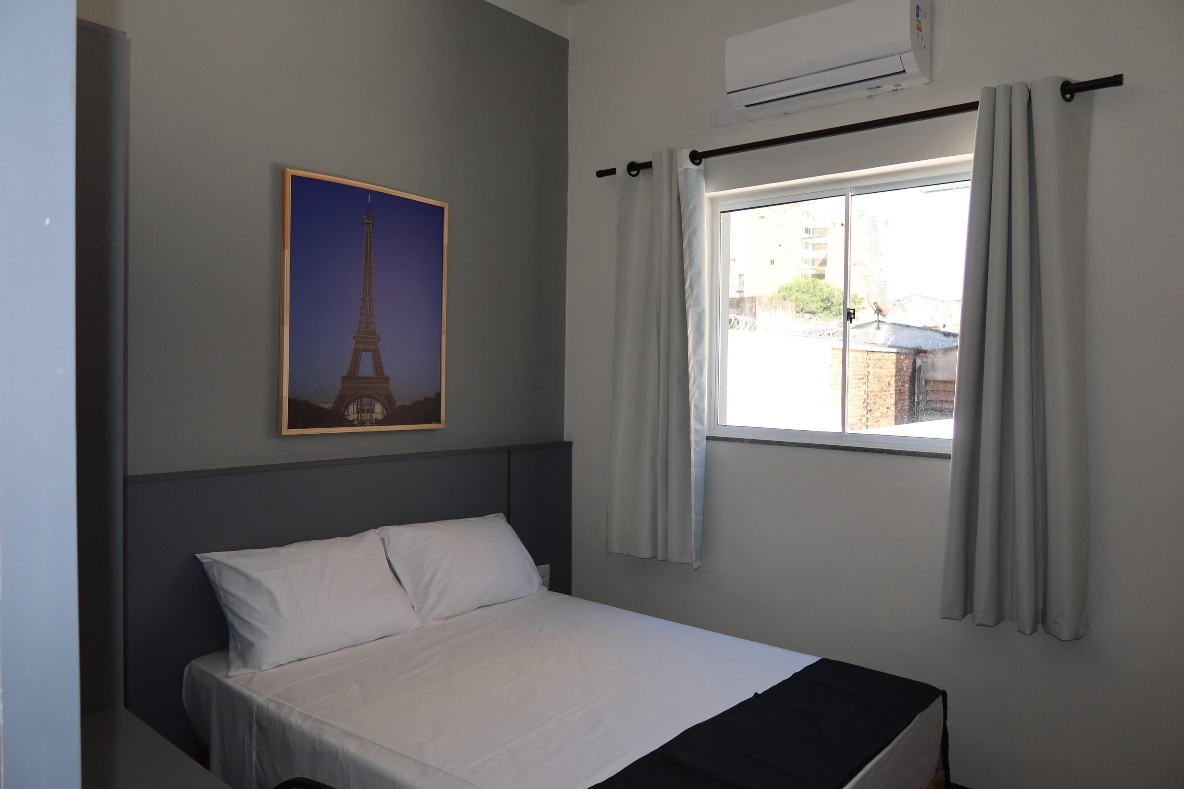 Une chambre à coucher avec un lit, une fenêtre avec des rideaux, un appareil de climatisation et un tableau encadré représentant la Tour Eiffel.
