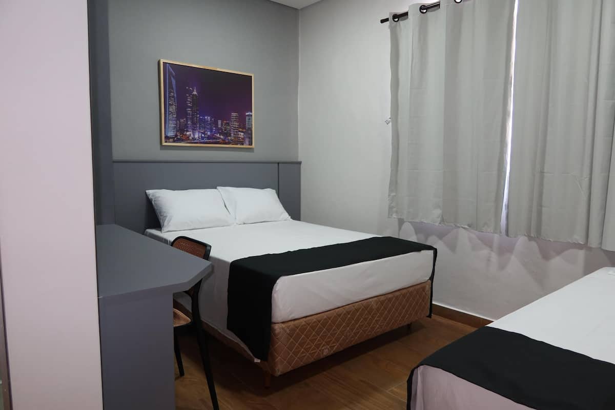Une chambre d’hôtel avec un lit, un bureau, une chaise, une fenêtre avec des rideaux et un tableau représentant un paysage urbain accroché au mur.