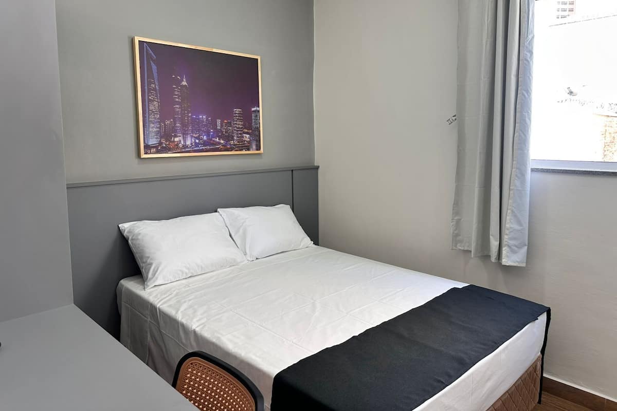 Une chambre à coucher avec un lit, une chaise, une fenêtre avec des rideaux et un tableau représentant un paysage urbain accroché au mur.