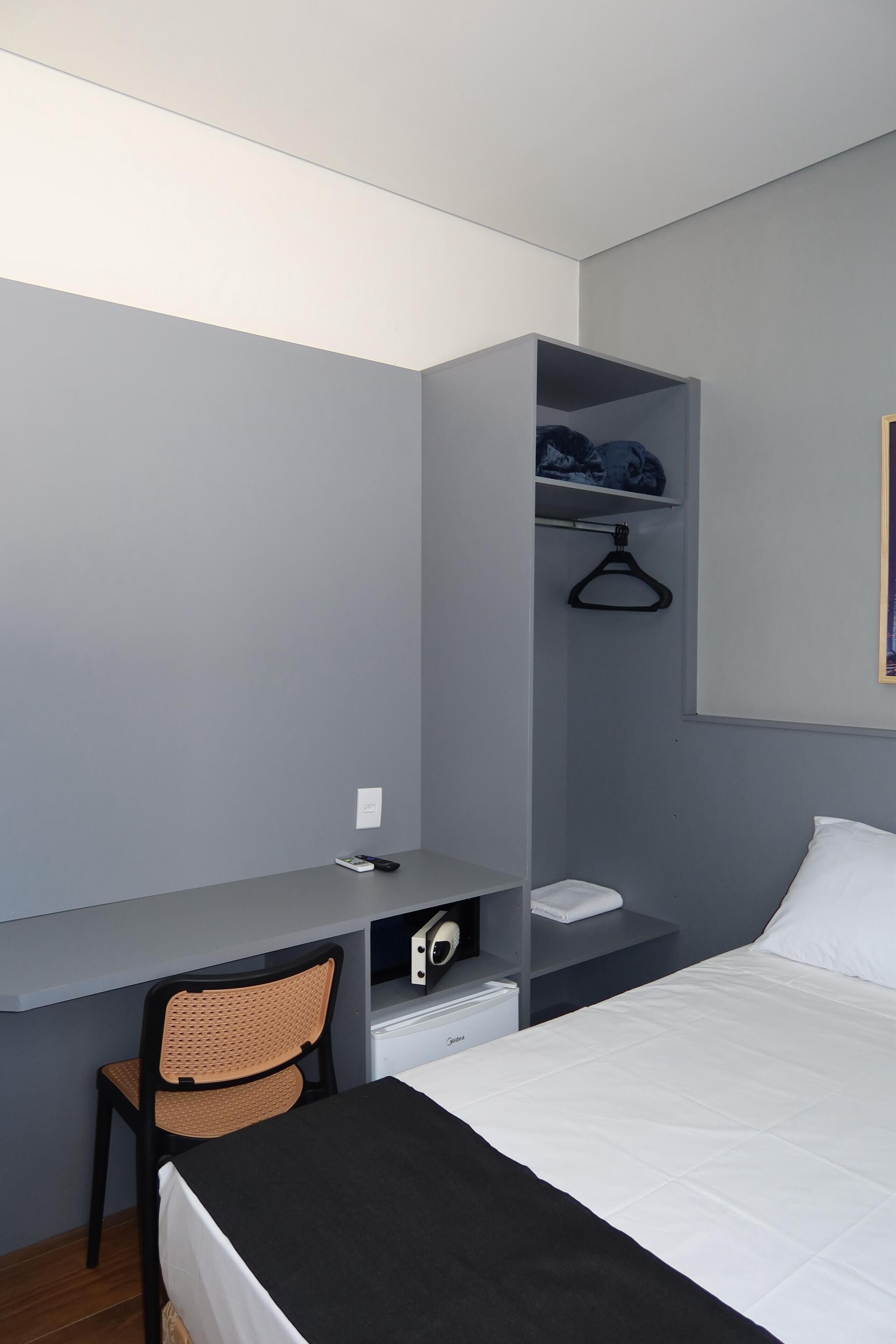A bedroom with a bed, a desk, a chair, a hanging clothes rack, and a small shelf with a towel.