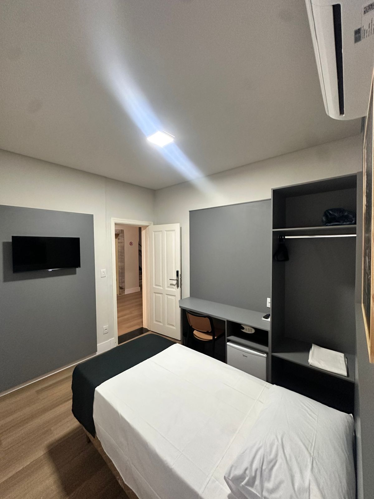 Quarto de hotel com cama, escrivaninha, cadeira, televisão e guarda-roupa.