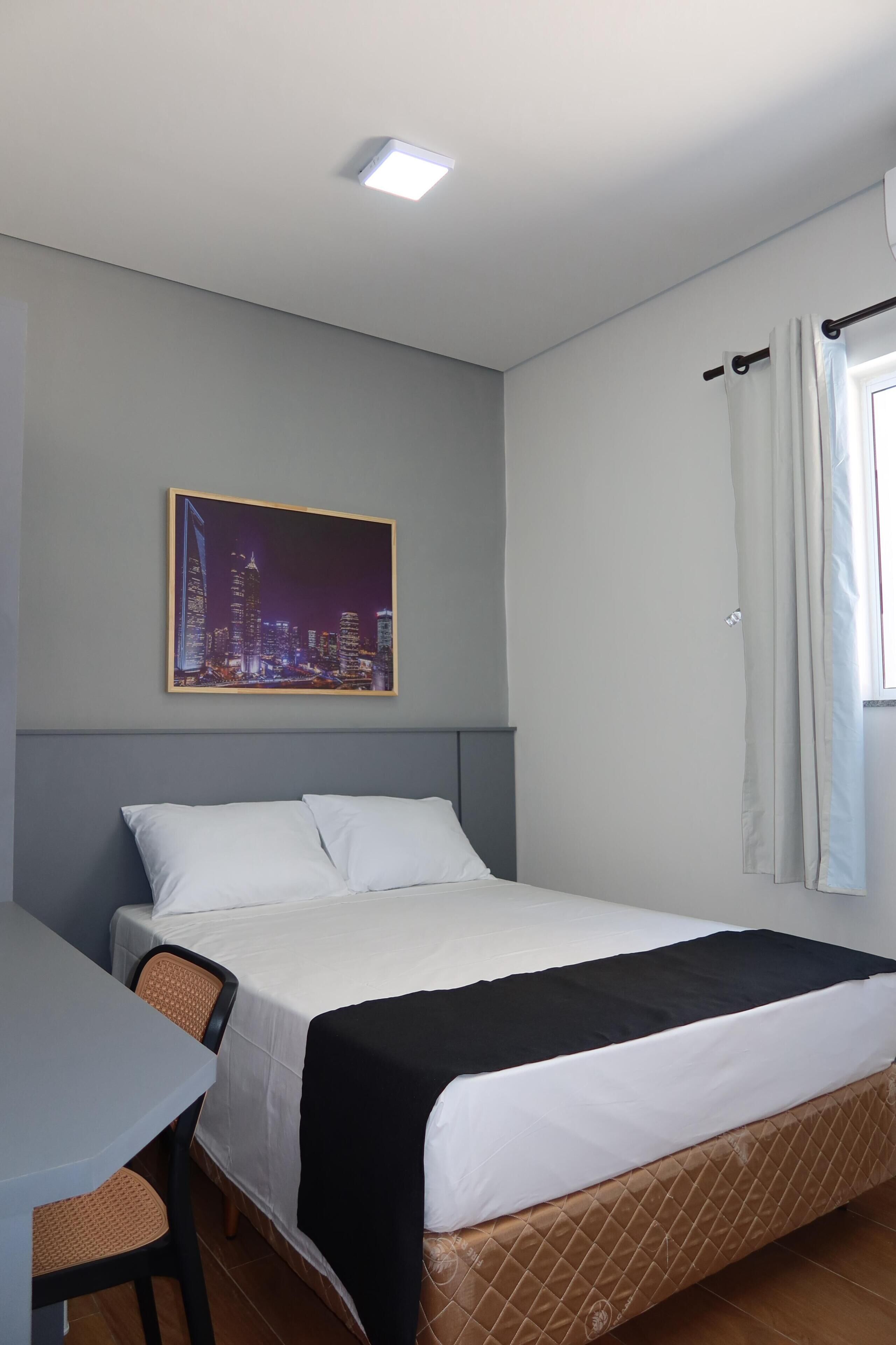 Quarto de hotel com cama, escrivaninha, cadeira, pintura de paisagem urbana e janela com cortinas.