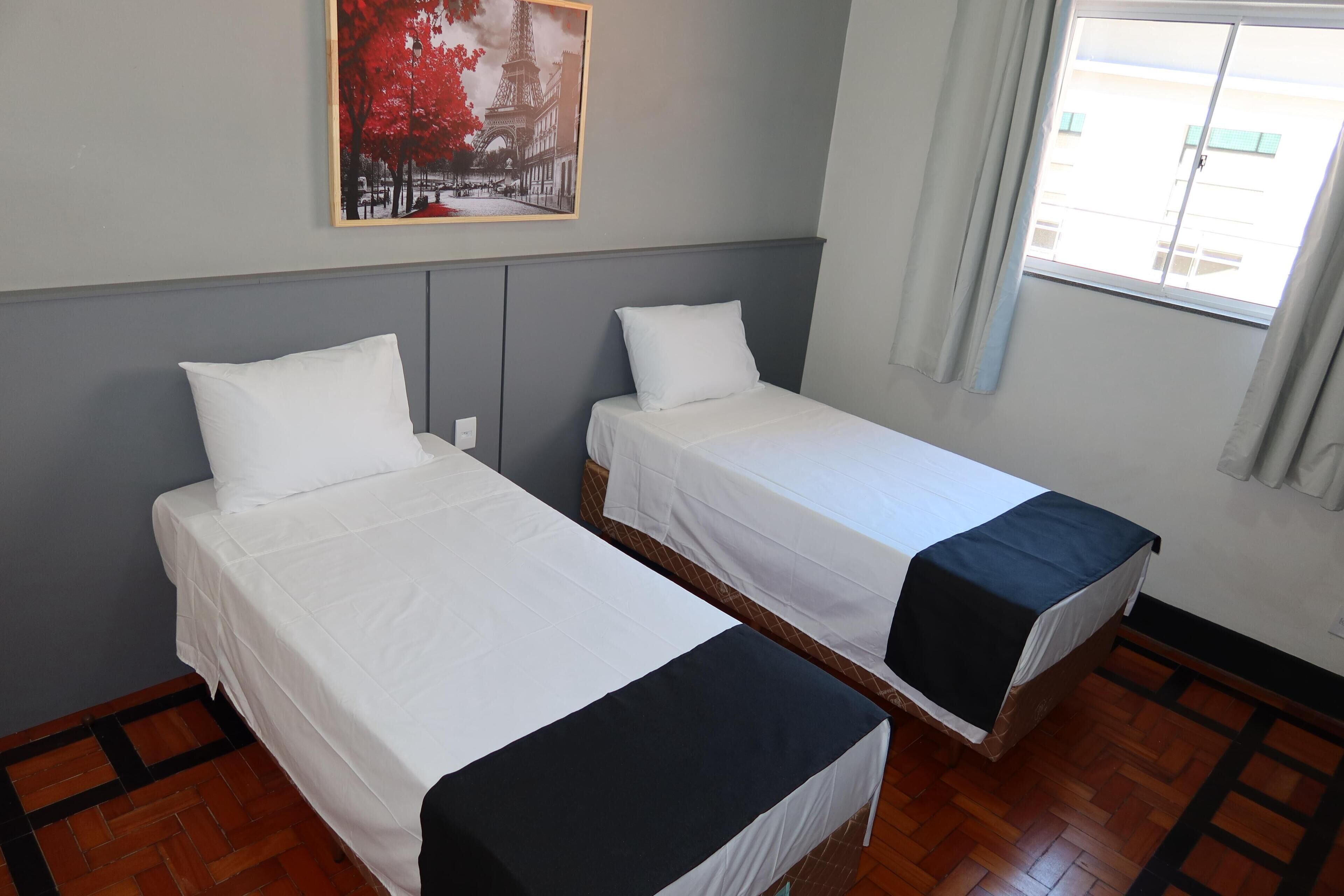 A hotel room with two single beds, a window with curtains, a wooden floor, and a painting on the wall.