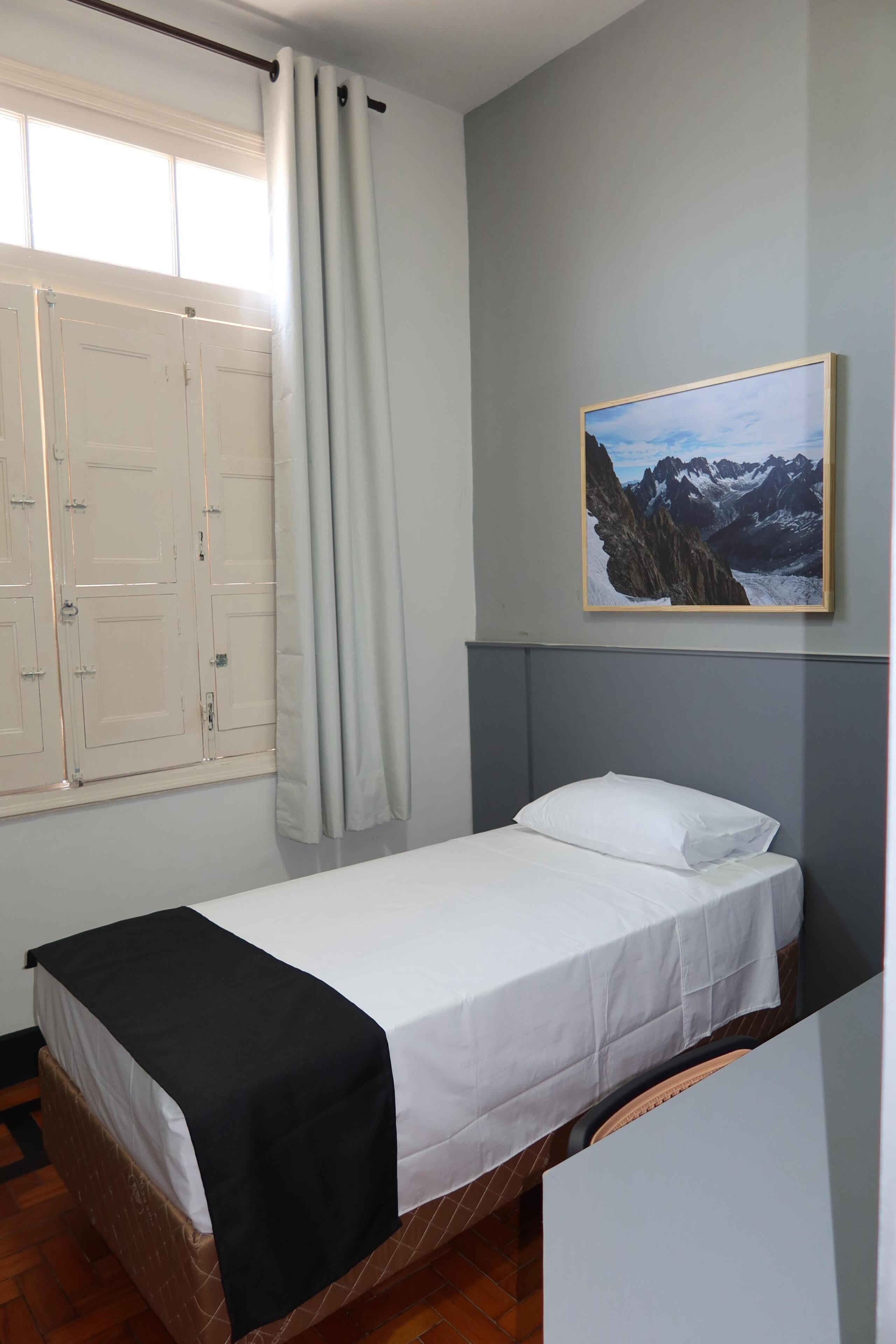 Uma cama de solteiro com colchão branco e manta preta, uma janela com cortinas brancas e um quadro com paisagem de montanhas na parede.