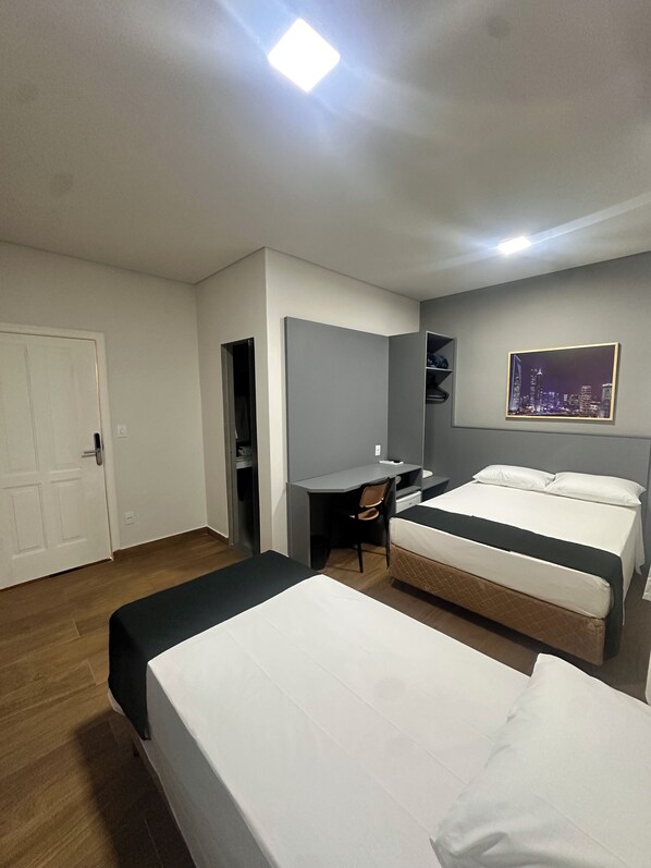Une chambre d’hôtel avec deux lits, un bureau, une chaise, une armoire et un tableau représentant un paysage urbain accroché au mur.