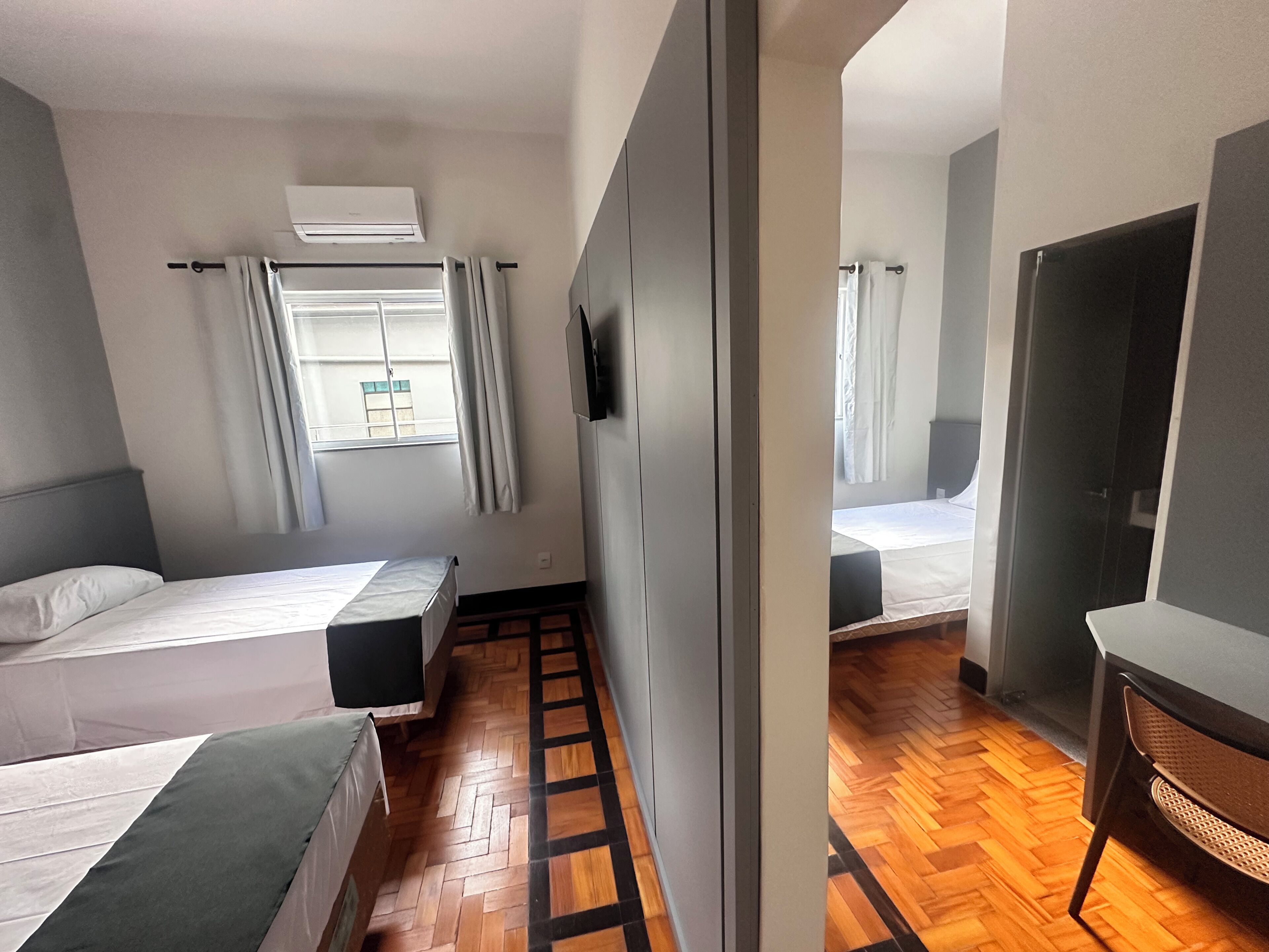 Quarto de hotel com duas camas, uma janela com cortinas, piso de madeira e um guarda-roupa.