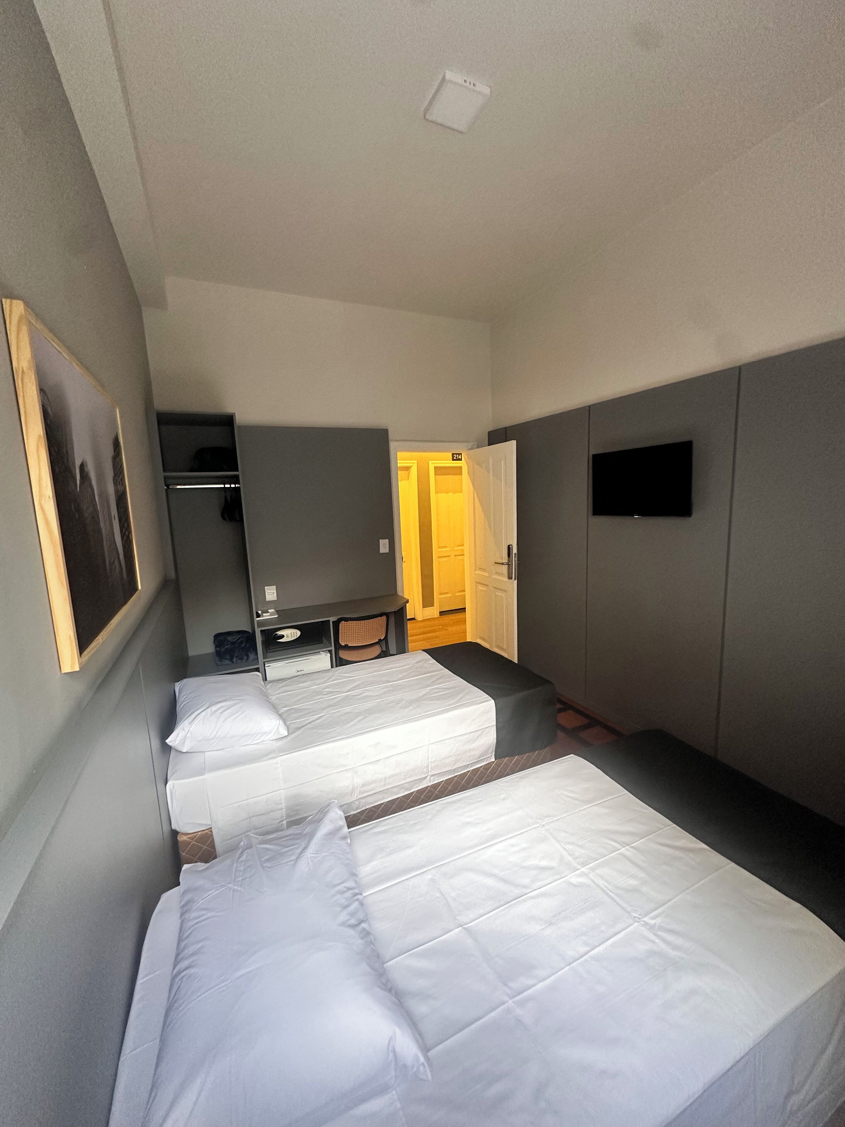 Quarto de hotel com cama, escrivaninha, cadeira, televisão e guarda-roupa.