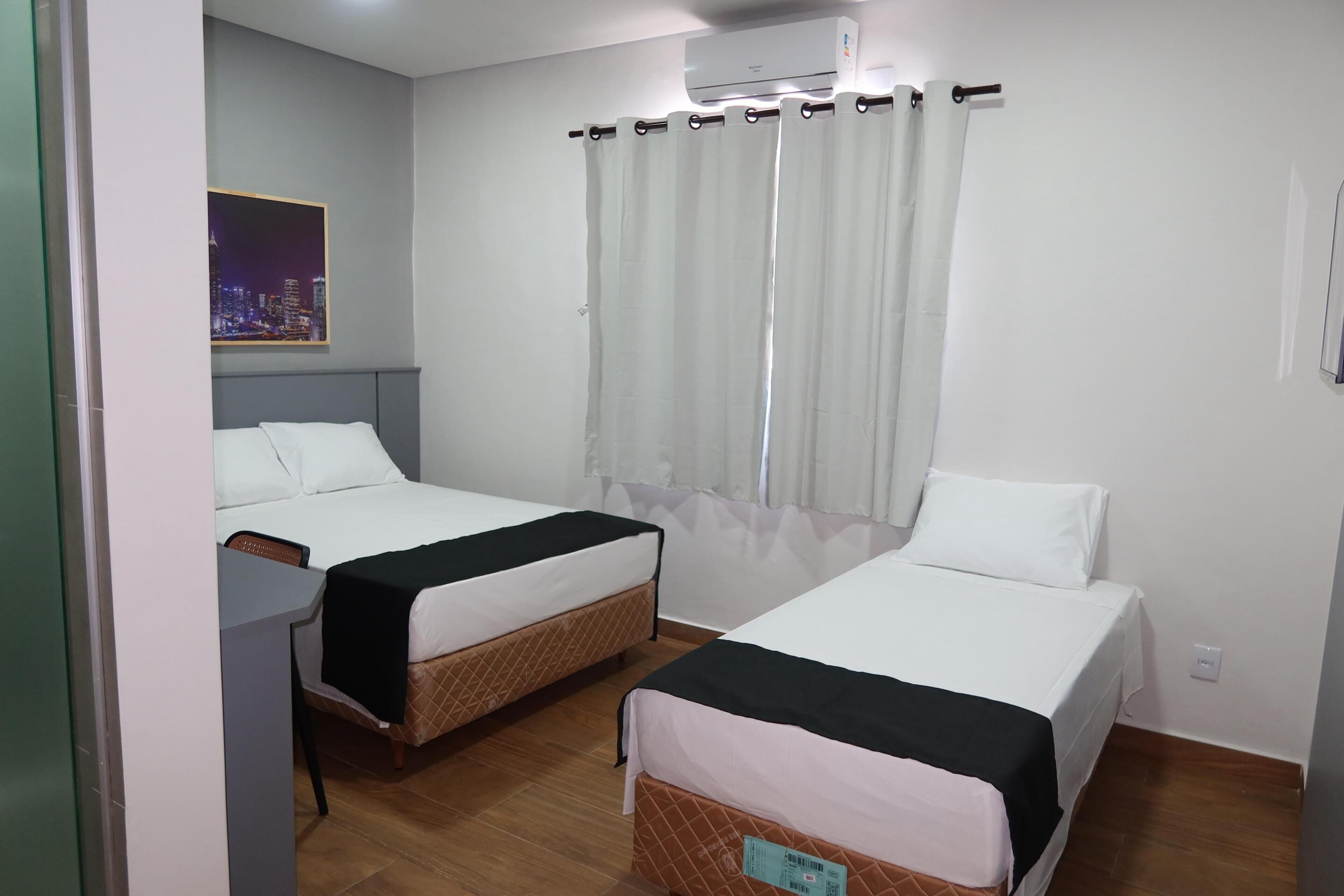A hotel room with two beds, a desk, a window with curtains, and a cityscape view.