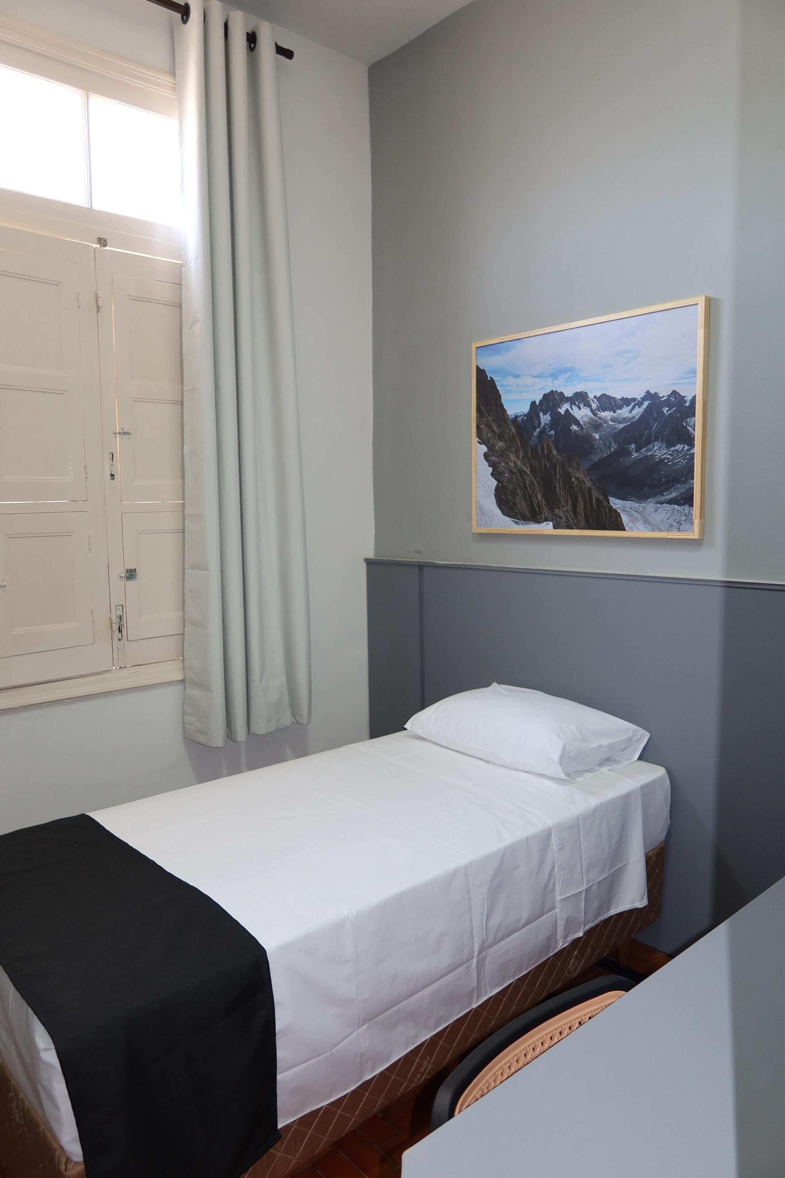 Un letto singolo con materasso e cuscino bianchi, una coperta nera, una finestra con tende e una cornice con un'immagine di montagne.