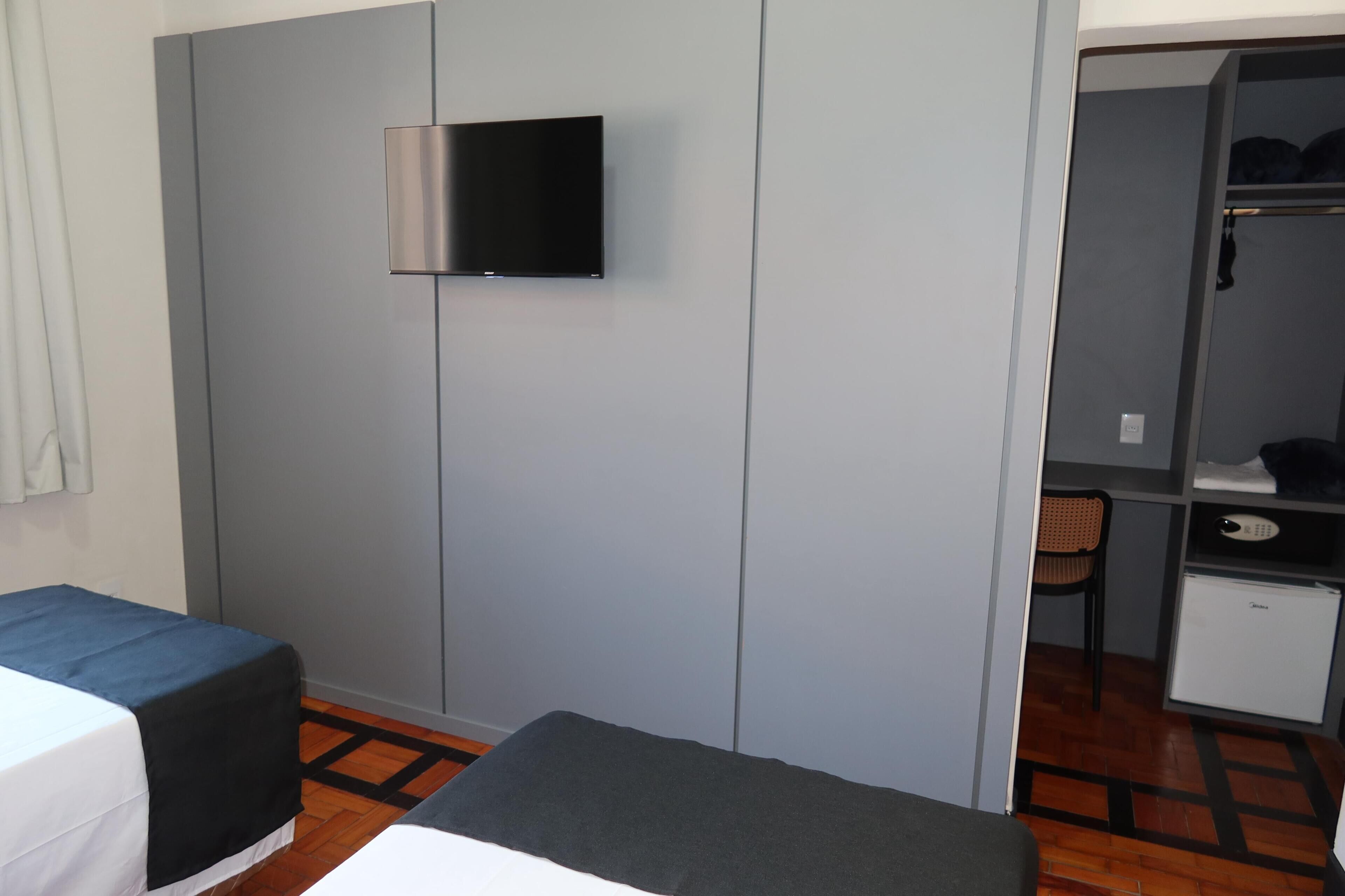 Una habitación con una cama, un escritorio pequeño, una silla, un televisor montado en la pared y un armario.