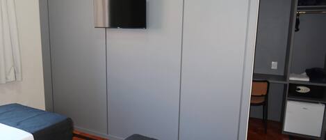 Una habitación con una cama, un escritorio pequeño, una silla, un televisor montado en la pared y un armario.
