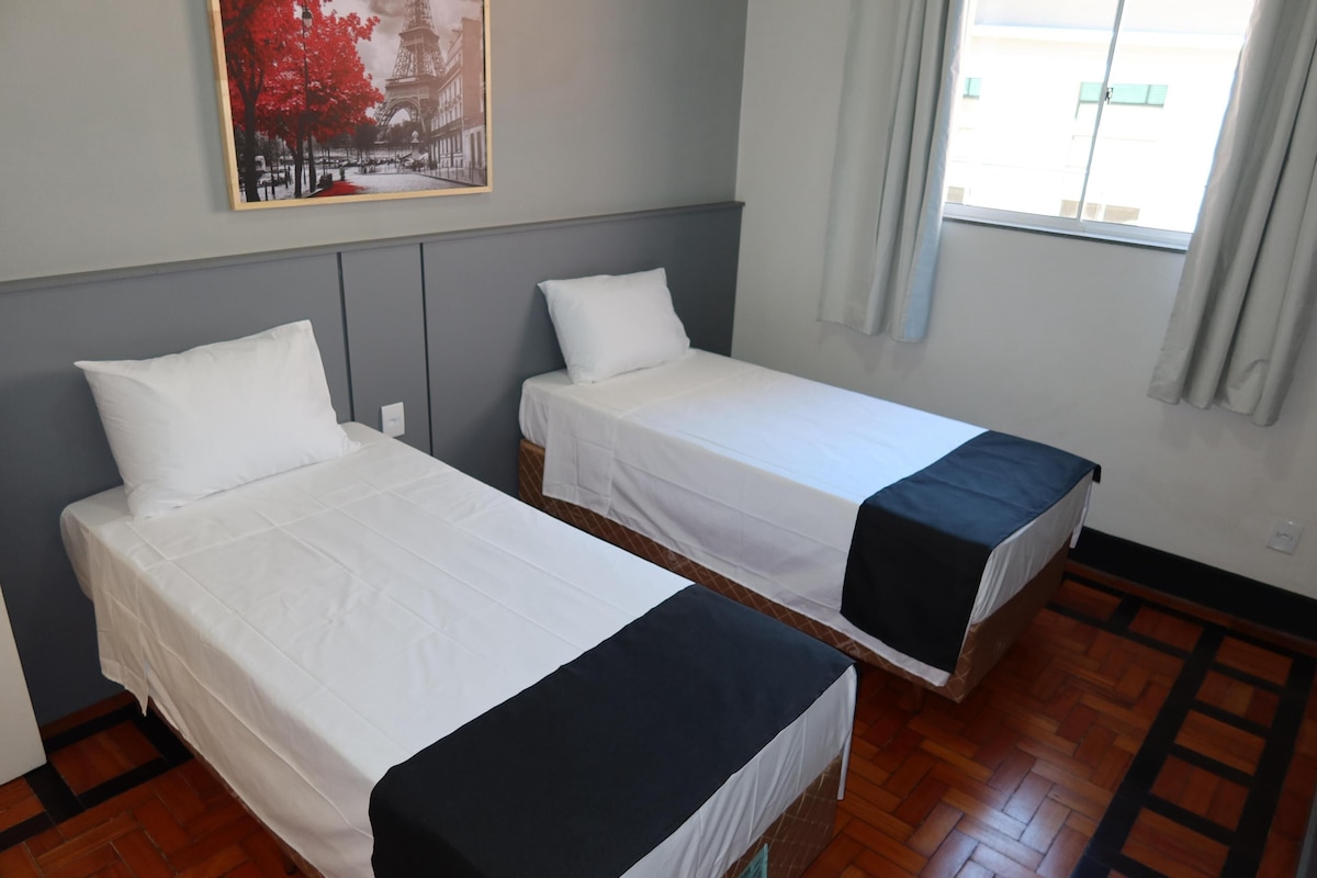 A room with two beds, a painting of the Eiffel Tower, a window with curtains, and wooden flooring.