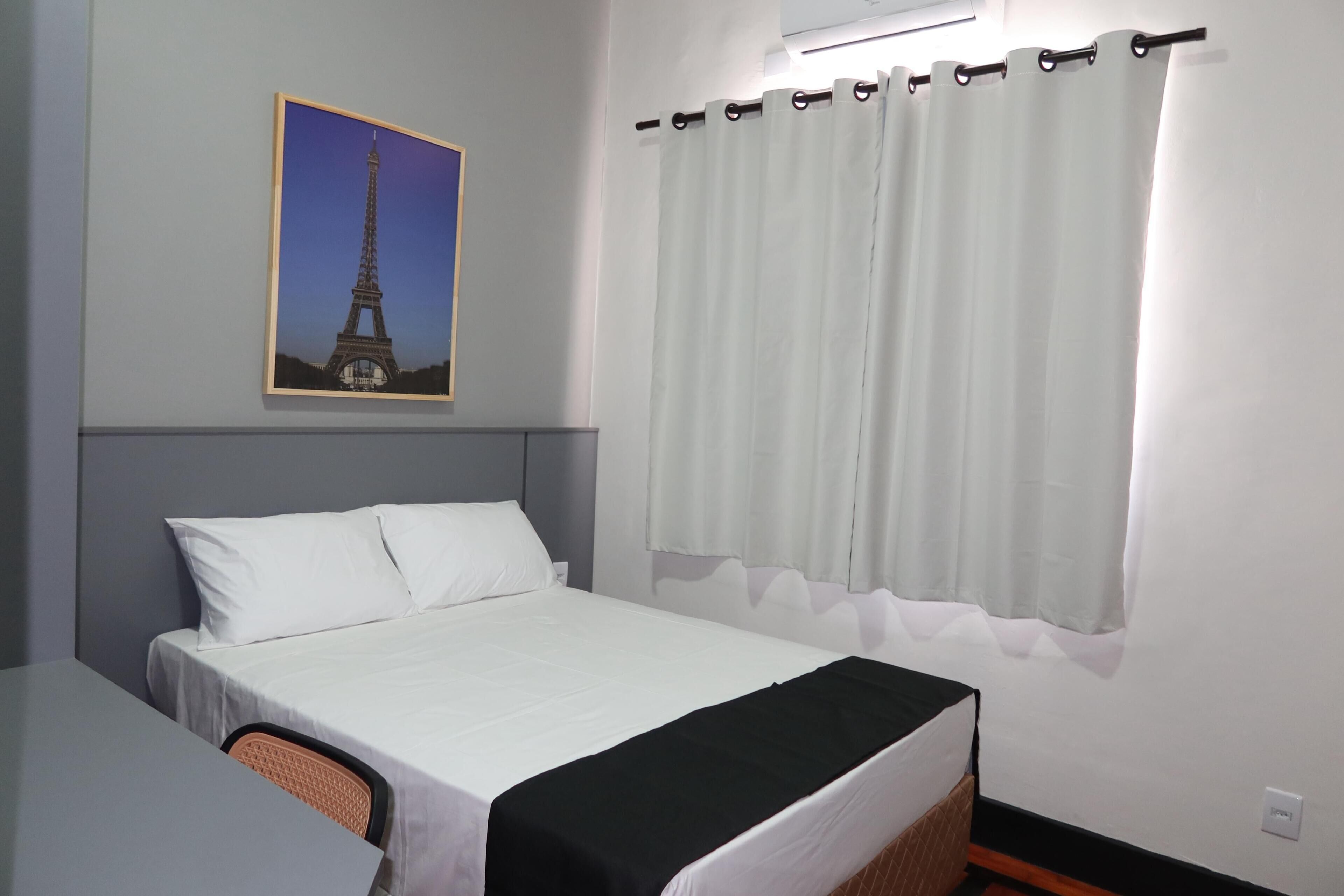 Una habitación de hotel con cama, escritorio, silla, aire acondicionado de pared y cortina.