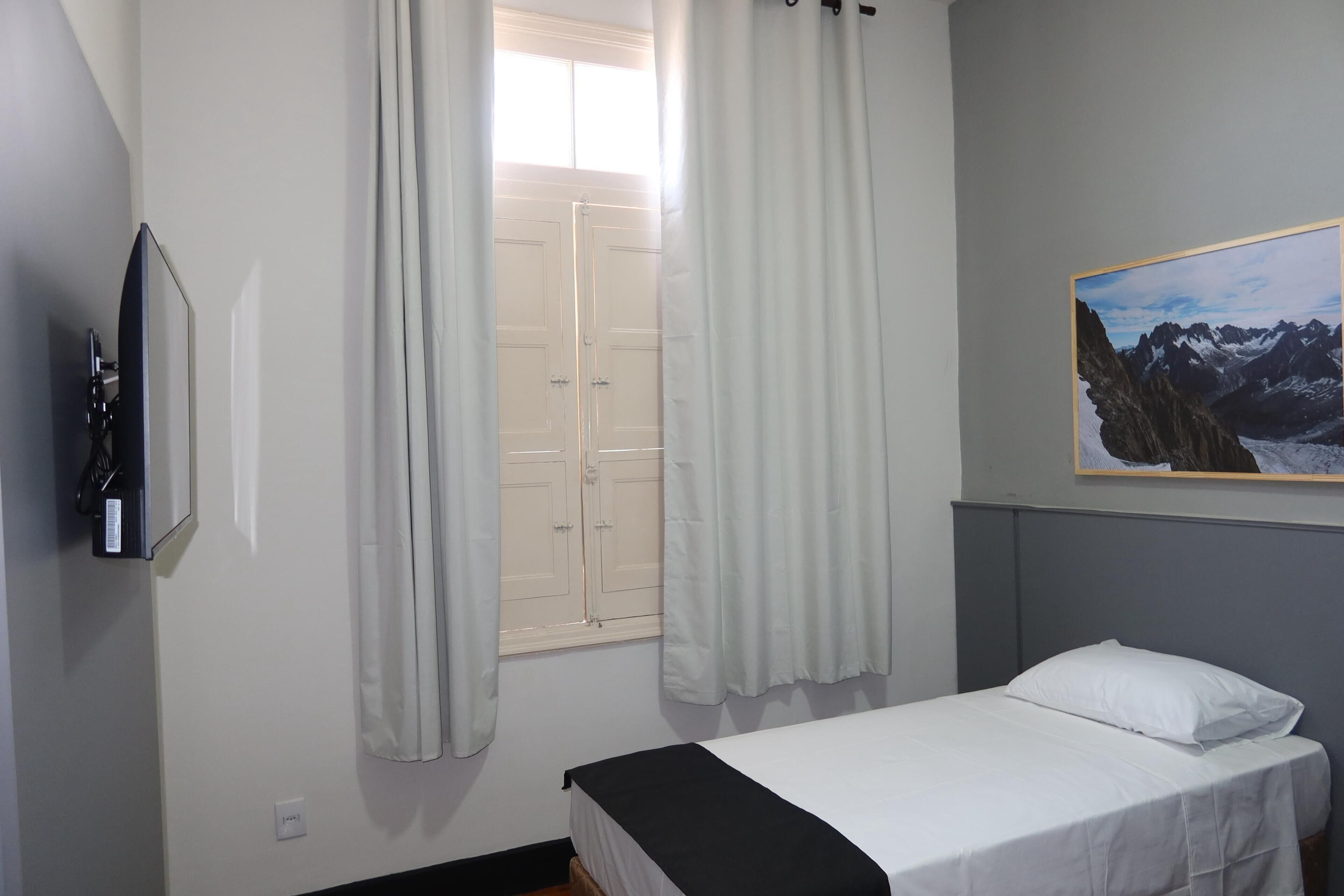 Quarto com uma cama, televisão presa na parede, janela com cortinas e um quadro pendurado na parede.