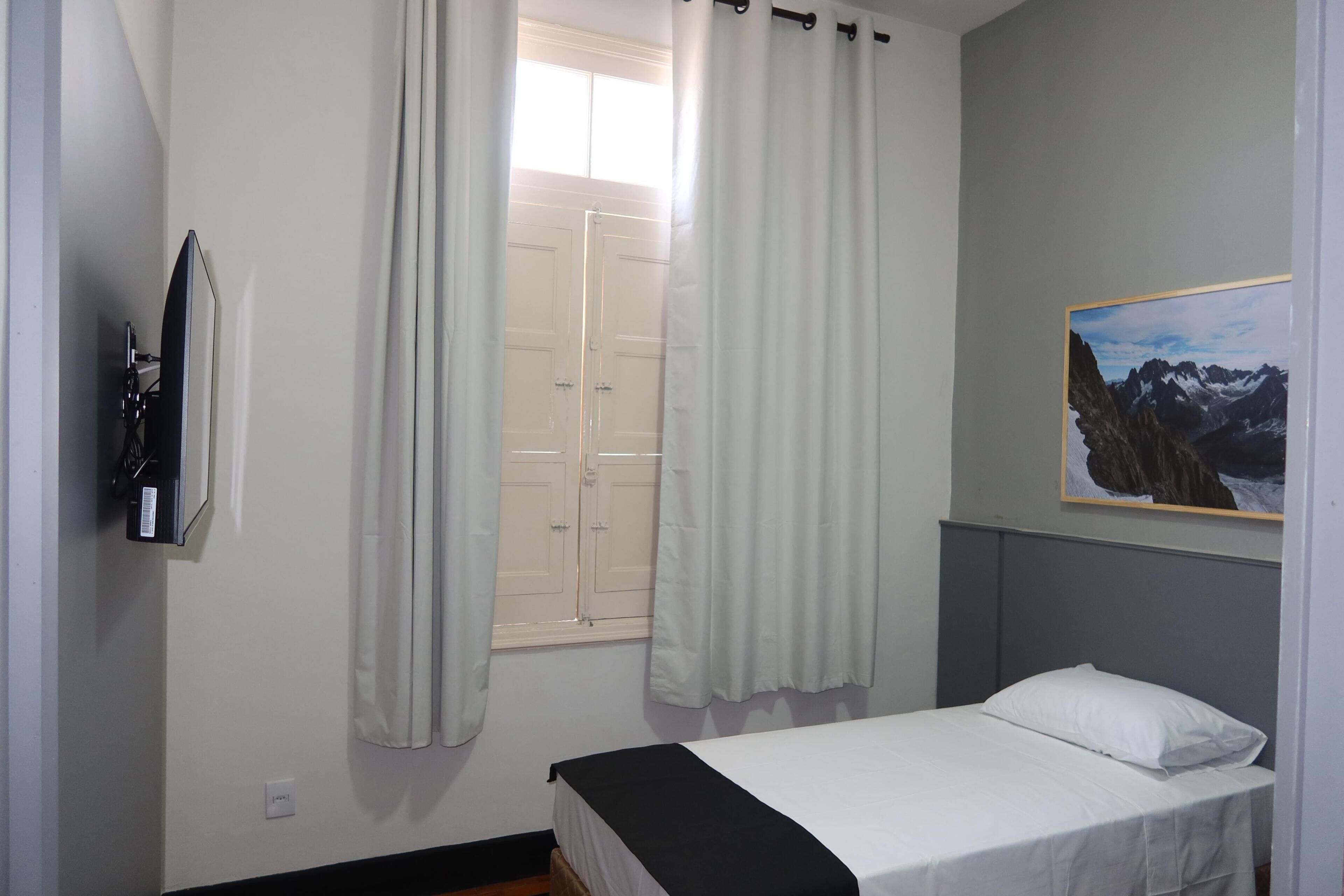 A bedroom with a bed, a TV mounted on the wall, a window with curtains, and a framed picture on the wall.