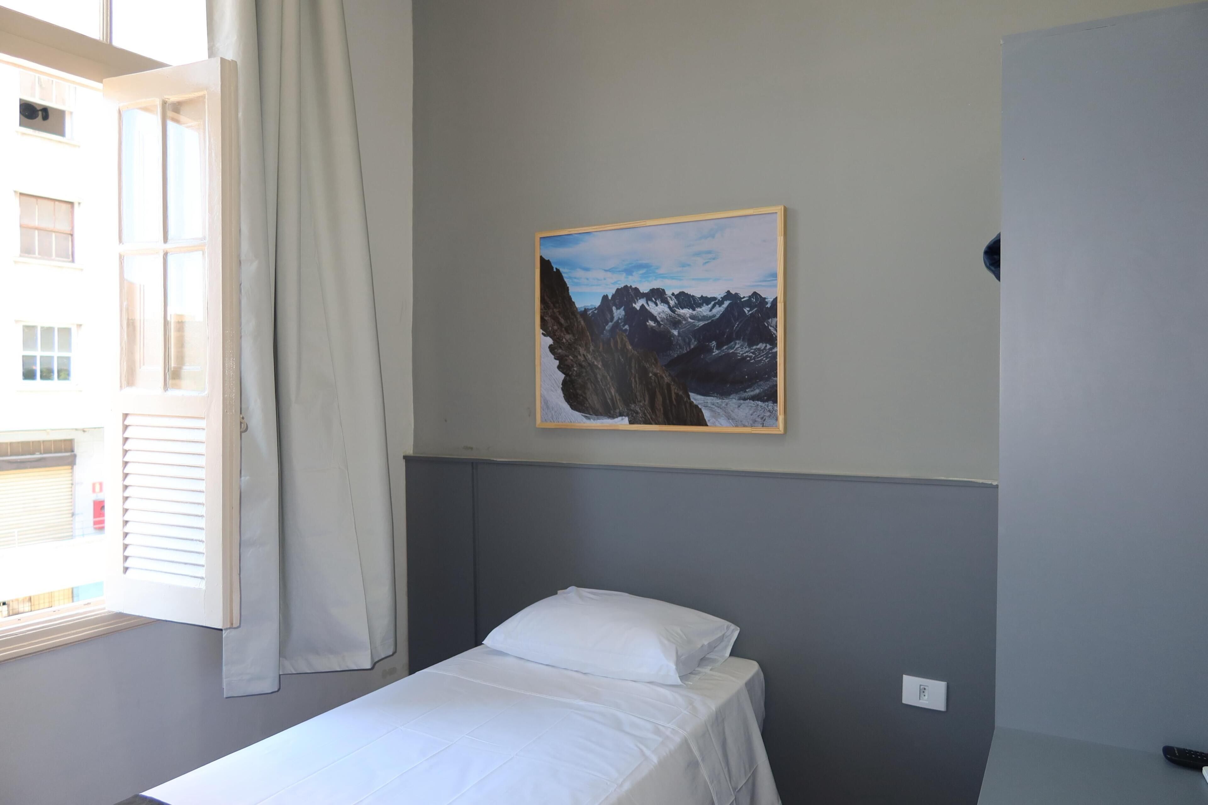 Quarto de hotel com cama, janela com cortinas, um quadro de montanhas na parede e controle remoto no chão.