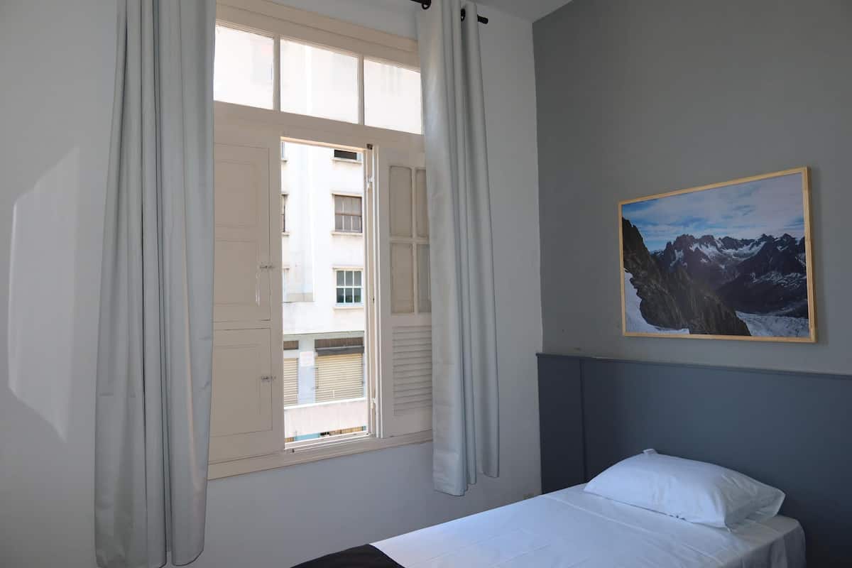 Une chambre à coucher avec un lit, une fenêtre avec des rideaux, un tableau accroché au mur et une vue sur les bâtiments à l’extérieur.