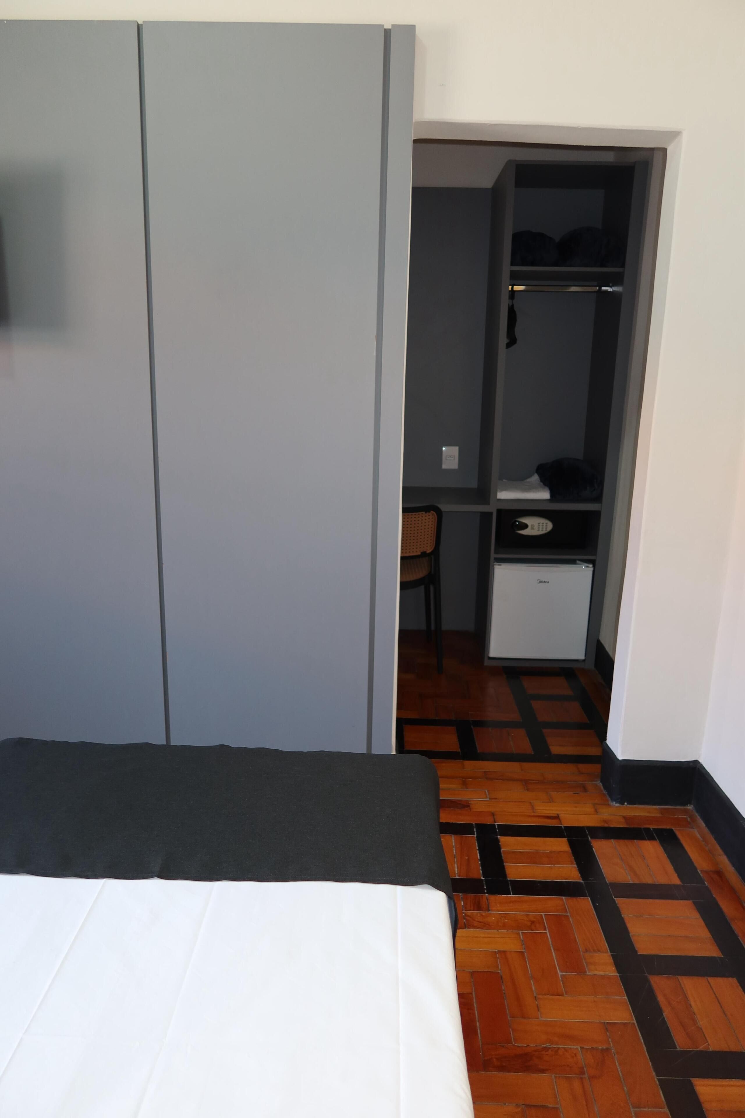 A bedroom with a bed, a wardrobe, a desk with a chair, and a small refrigerator.