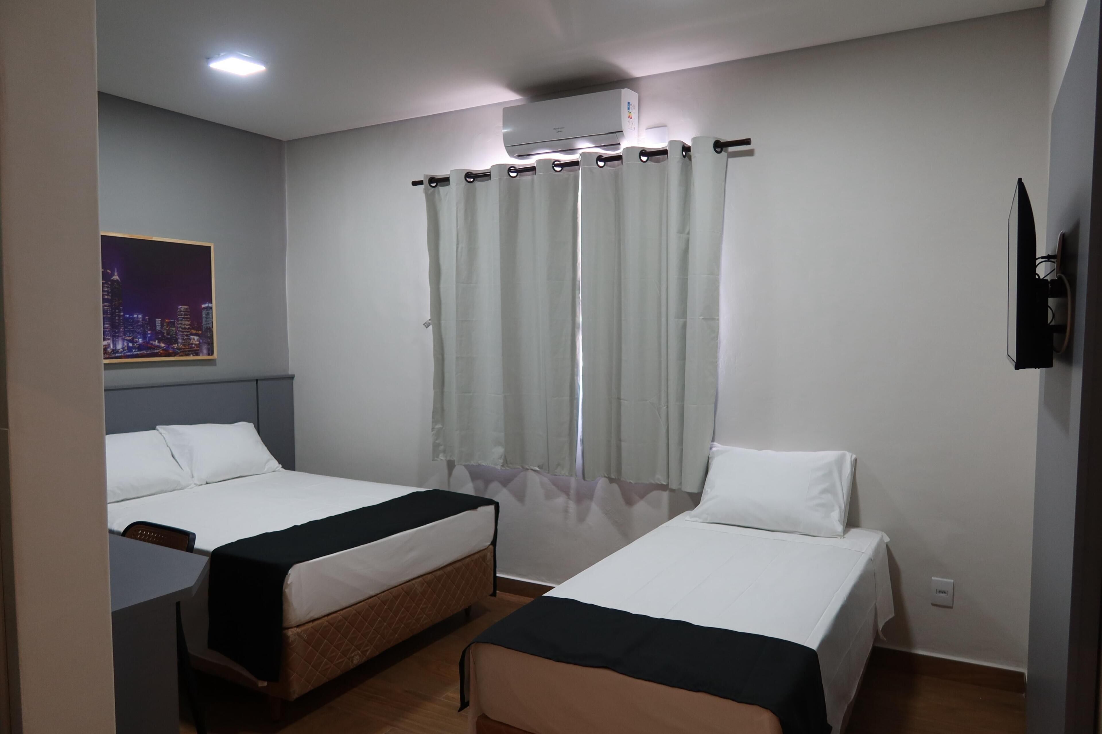 A hotel room with two beds, a desk, an air conditioning unit, and a cityscape poster.