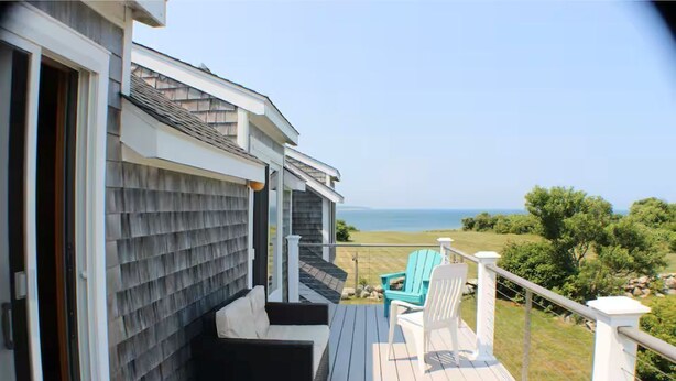 A deck with outdoor furniture, a view of the ocean, and a house with shingled siding. - Relaxing Spring Street Cottage with Ocean Views (New Shoreham)