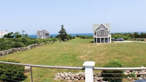 A coastal property with a stone wall, a large house with a porch, and a view of the ocean. - Relaxing Spring Street Cottage with Ocean Views (New Shoreham)