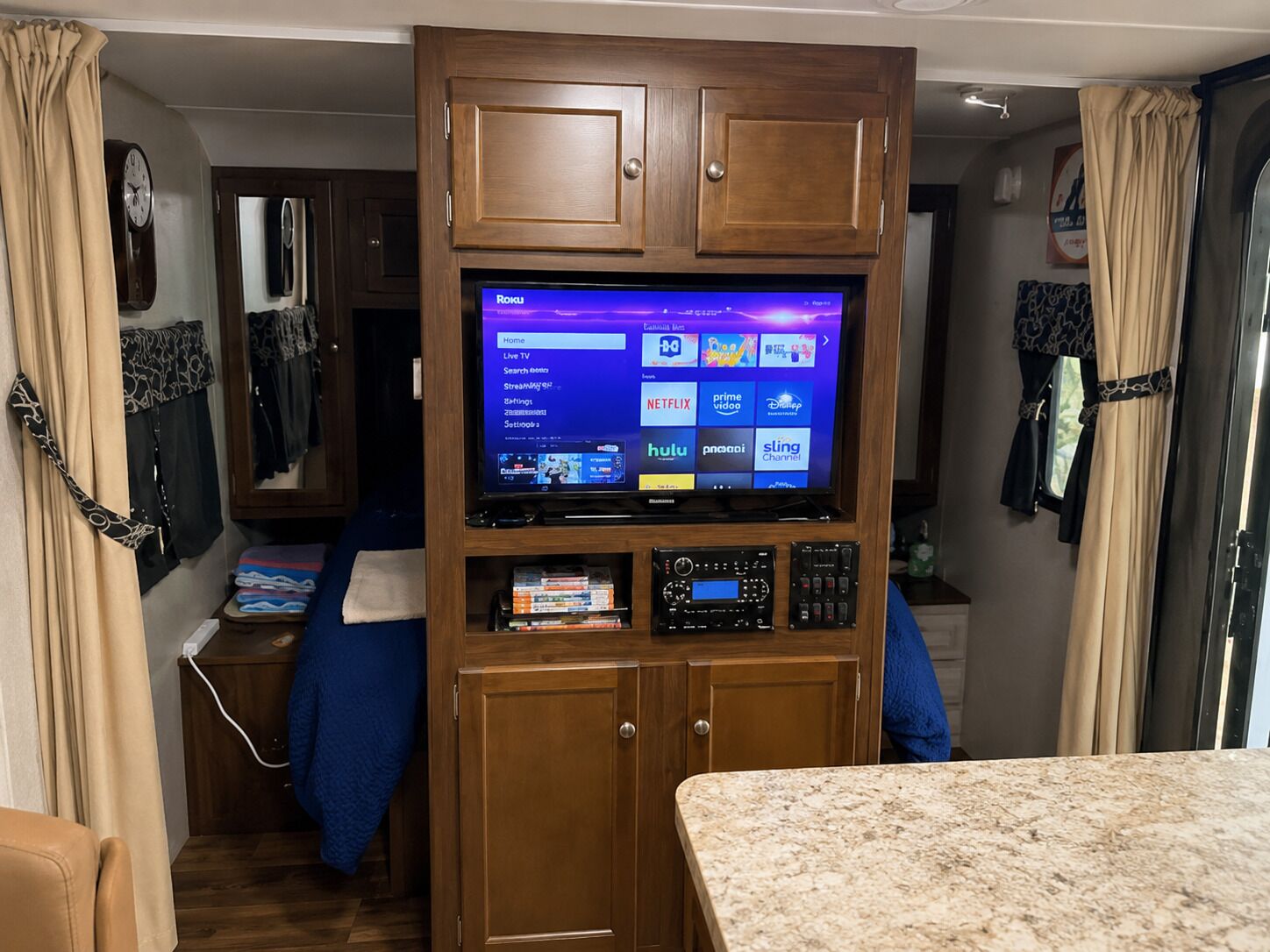 A compact RV interior with a television mounted on a wooden cabinet, a clock, and a small kitchenette area.