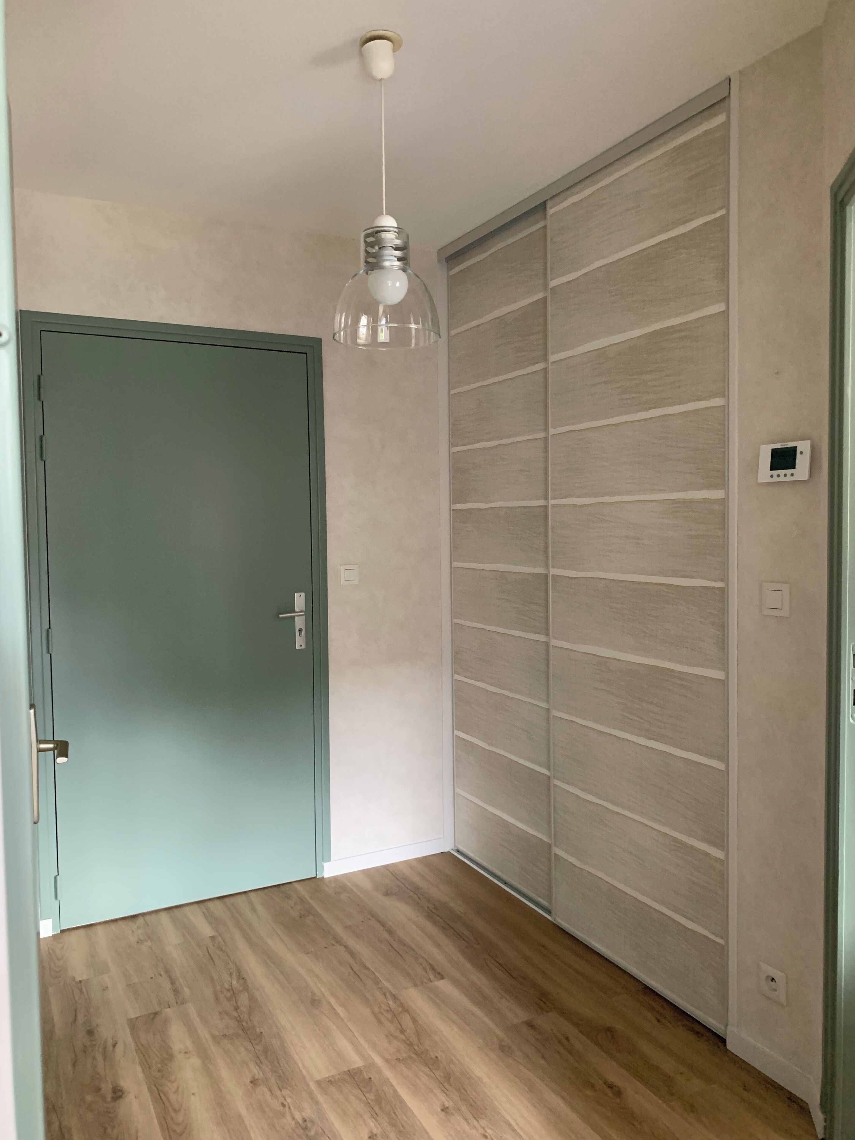 Une pièce avec un plancher en bois, une porte verte, un ventilateur de plafond et un thermostat fixé au mur.