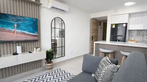 Ein modernes Wohnzimmer mit einer grauen Couch, einem großen Fernseher und einer Küchenzeile mit weißen Schränken und einer gefliesten Spritzschutzwand.
