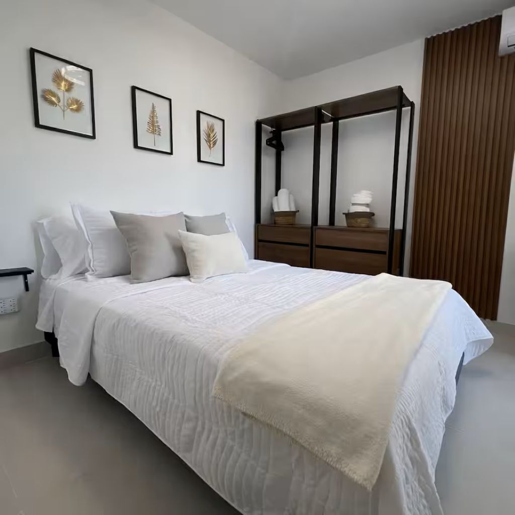 A neatly made bed with a white comforter and pillows, a wooden nightstand with a lamp, and a dresser with a mirror.