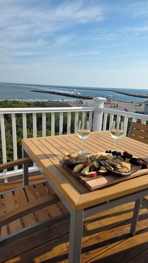 A wooden deck with a table set for two, overlooking a beach with a rocky breakwater.
