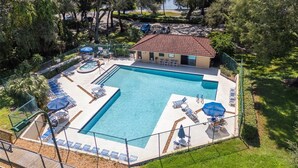 A swimming pool with lounge chairs and umbrellas, a small building with a red roof, and a fenced area with trees. - Beautiful Townhome/Fully Furnished in the heart of Palm Harbor. (Palm Harbor)