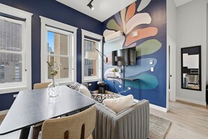 A modern living room with a dining area, large windows, and a colorful abstract wall mural.