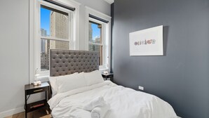 Quarto com cabeceira estofada, cama com roupa de cama branca, mesa de cabeceira com abajur e quadro pendurado na parede.