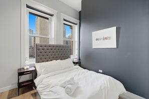 A bedroom with a tufted headboard, a bed with white linens, a nightstand with a lamp, and a framed picture on the wall.