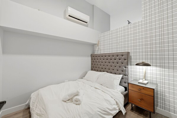 A bedroom with a bed, a nightstand, a lamp, an air conditioner, and a grid-patterned wall.