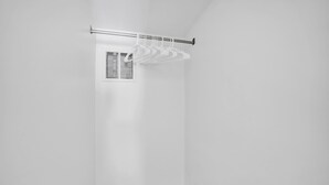 Esquina de una habitación con pared blanca, una barra de cortina metálica y perchas blancas, y un respiradero.