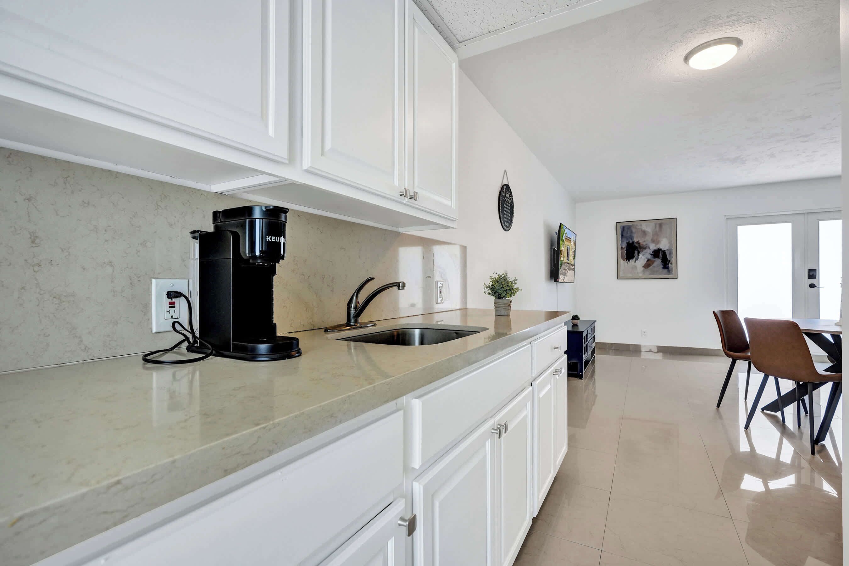 A modern kitchen with white cabinets, a coffee machine, a sink, and a dining area with chairs and a table.