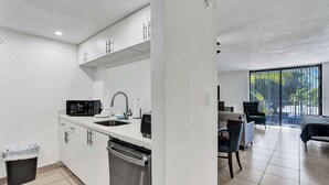 Een moderne keuken met witte kastjes, een vaatwasser en een magnetron. Er is een eethoek met een stoel en een tafel.
